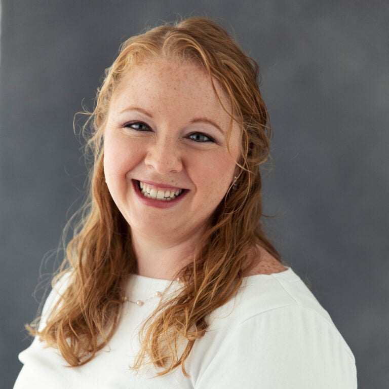 Freckled woman with red hair smiling, wearing a white shirt and pearl necklace.
