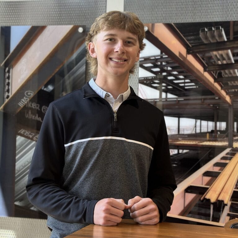 Young man with a mullet smiles, wearing a black and gray quarter-zip shirt, standing in front of a construction site mural.